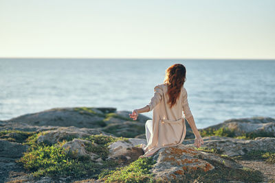 Woman sitting on rock by sea against clear sky