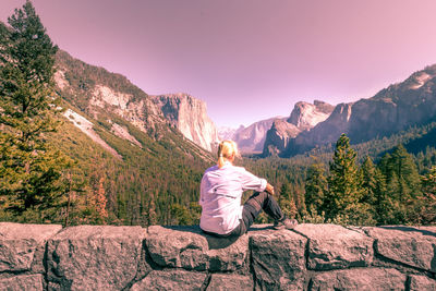 Rear view of woman sitting on retaining wall against mountains