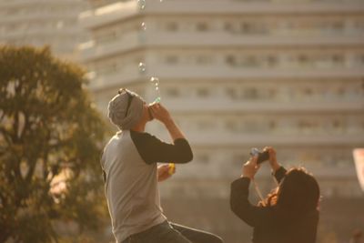 Man blowing bubbles by woman photographing against building