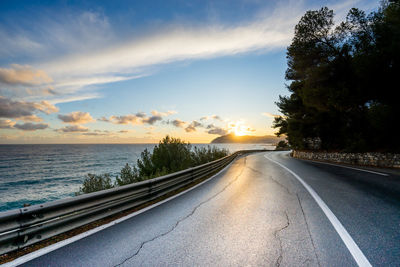 Road by trees against sky during sunset