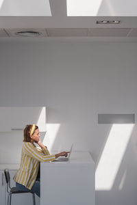 Busy pregnant female remote worker in stylish striped jacket and headband sitting at table and browsing laptop while working on project at home