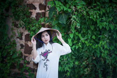 Young woman wearing asian style conical hat while standing against ivy