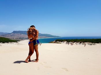 Side view of couple kissing while standing at beach against clear blue sky during sunny day