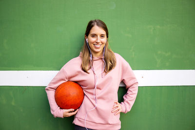 Portrait of smiling young woman holding basketball standing against wall