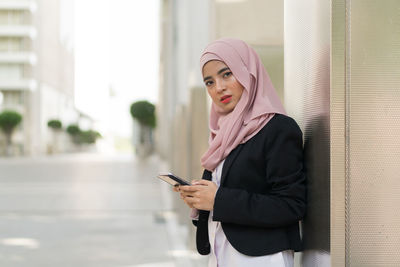 Portrait of businesswoman wearing hijab using mobile phone while standing against building