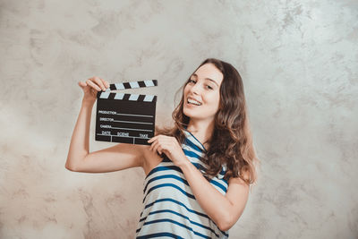 Portrait of cheerful young woman holding film slate by wall