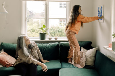 Girl arranging painting on wall while grandmother sitting in sofa at home