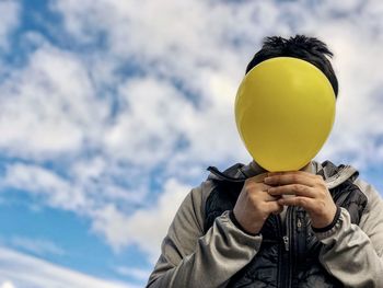Low angle view of man holding yellow balloon against cloudy sky.