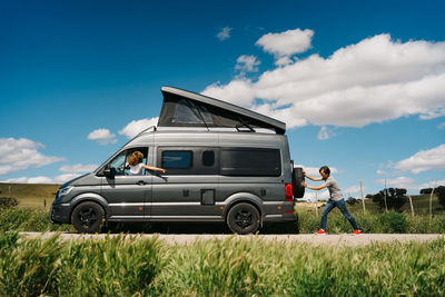 Side view of young man pushing camper van while woman sitting at steering wheel as having troubles during trip in summer countryside