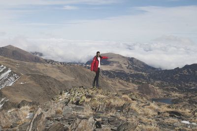Young man with red jacket standing on mountain against sky.