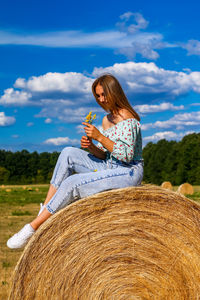 Young woman sitting on hay against sky