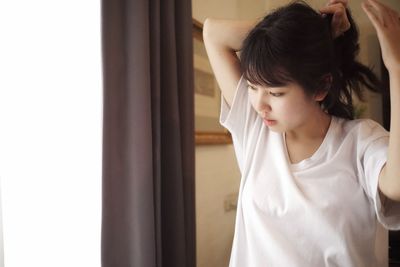 Young woman tying hair while standing by window at home