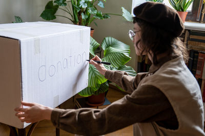 Interested girl altruist student volunteer preparing box for donations writing inscription at home.