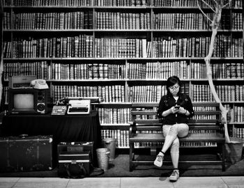 Woman using mobile phone while sitting on bench against bookshelf