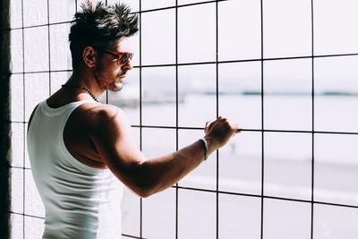 Side view of young man standing against window