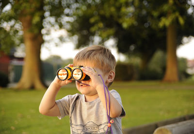 Boy looking through binoculars while sitting against trees in park