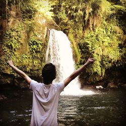 Rear view of teenage boy with arms outstretched standing against waterfall