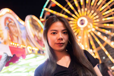 Portrait of young woman with illuminated carousel in amusement park