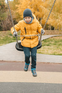 Happy boy in a hat and a yellow jacket, riding on a swing, on the street. 