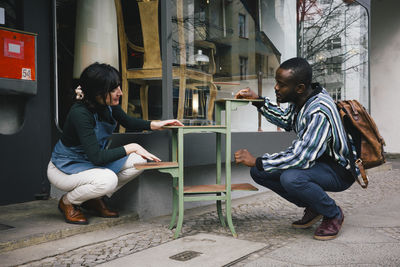 Female upcycling store owner showing furniture to customer while crouching at sidewalk