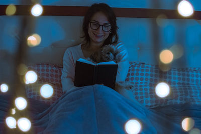Young woman with dog reading book while sitting on bed at home