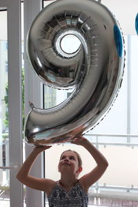 Girl holding number 9 shape balloon against glass door
