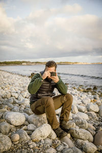 Man photographing from camera while sitting on rock at beach against sky
