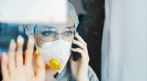 Woman talking over phone while wearing protective workwear seen through glass window