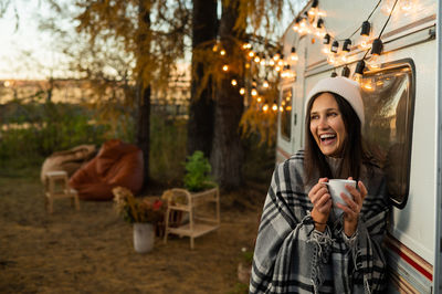 Caucasian woman in a knitted hat wrapped in a plaid and drinks a warming drink outdoors. travel in a