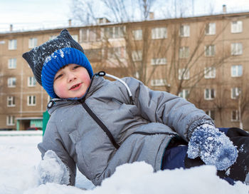 Boy wearing knit hat lying on snow covered land against building