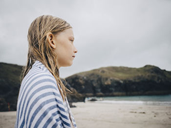 Side view of blond girl wrapped in striped blue towel standing at beach against sky