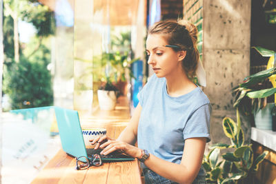 Stylish blonde hipster girl in a bright cafe at a table by the window typing in a laptop message. 