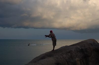 Side view of man fishing by sea against cloudy sky during sunset