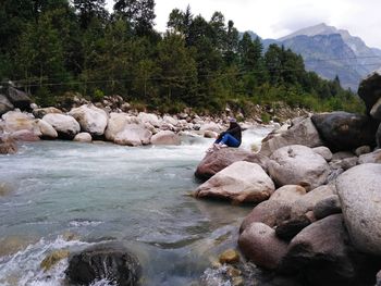 Man sitting on rock by river against sky