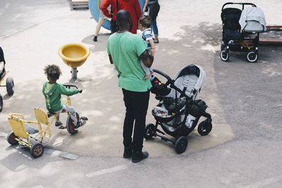 High angle view of father carrying toddler while standing by son riding tricycle at park