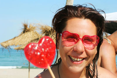 Portrait of smiling woman wearing heart shape sunglasses with lollipop at beach