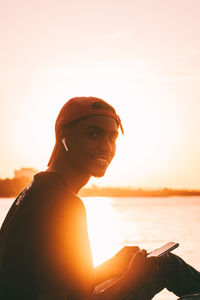 Side view of young man sitting at ocean using mobile phone against clear sky