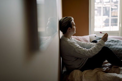 Side view of upset woman holding pregnancy test result sitting on bed at home