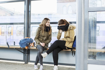 Full length of happy young woman looking at friend covering face with scarf at train station