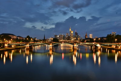 Illuminated bridge over river by buildings against sky at dusk