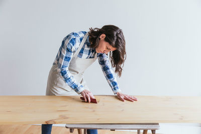 Focused skilled female woodworker with black hair in apron polishing wooden board with sandpaper while working in light professional workshop on white background
