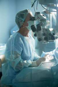 Adult female doctor in sterile mask and ornamental medical cap looking through surgical microscope against crop coworker in hospital