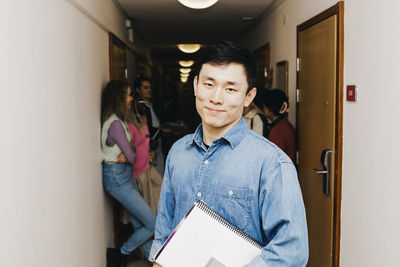 Confident young man with book while multiracial friends in background at corridor in dorm