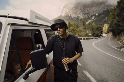 Young man wearing sunglasses standing next to van during road trip