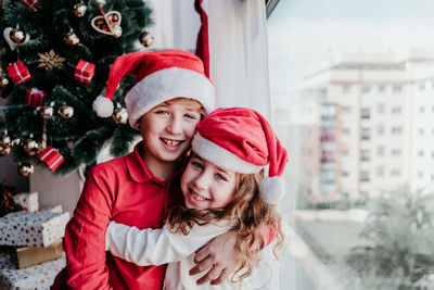 Portrait of siblings wearing santa hats embracing at home