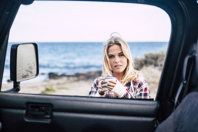 Portrait of young woman having drink seen through vehicle window at beach