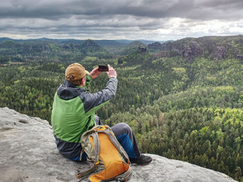 Ginger hair man takes photos with smart phone on rocky peak. rocky view point above forest valley.