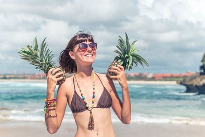 Happy young woman holding pineapples at beach against sky