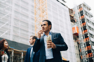 Low angle view of businessman eating wrap sandwich while standing by colleagues against building in city
