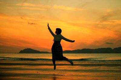 Woman jumping on shore at beach against sky during sunset